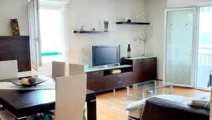 A modern living room with a dark wooden dining table, white chairs, a glass coffee table, a dark sofa, and a television on a wooden stand. - Cosy mountain view apartment, 2 bedrooms, terrace and parking box (Hendaye)