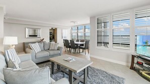 A modern living room with a grey sofa, a coffee table, and a TV. There is a dining area with a table and chairs, and large windows with blinds.