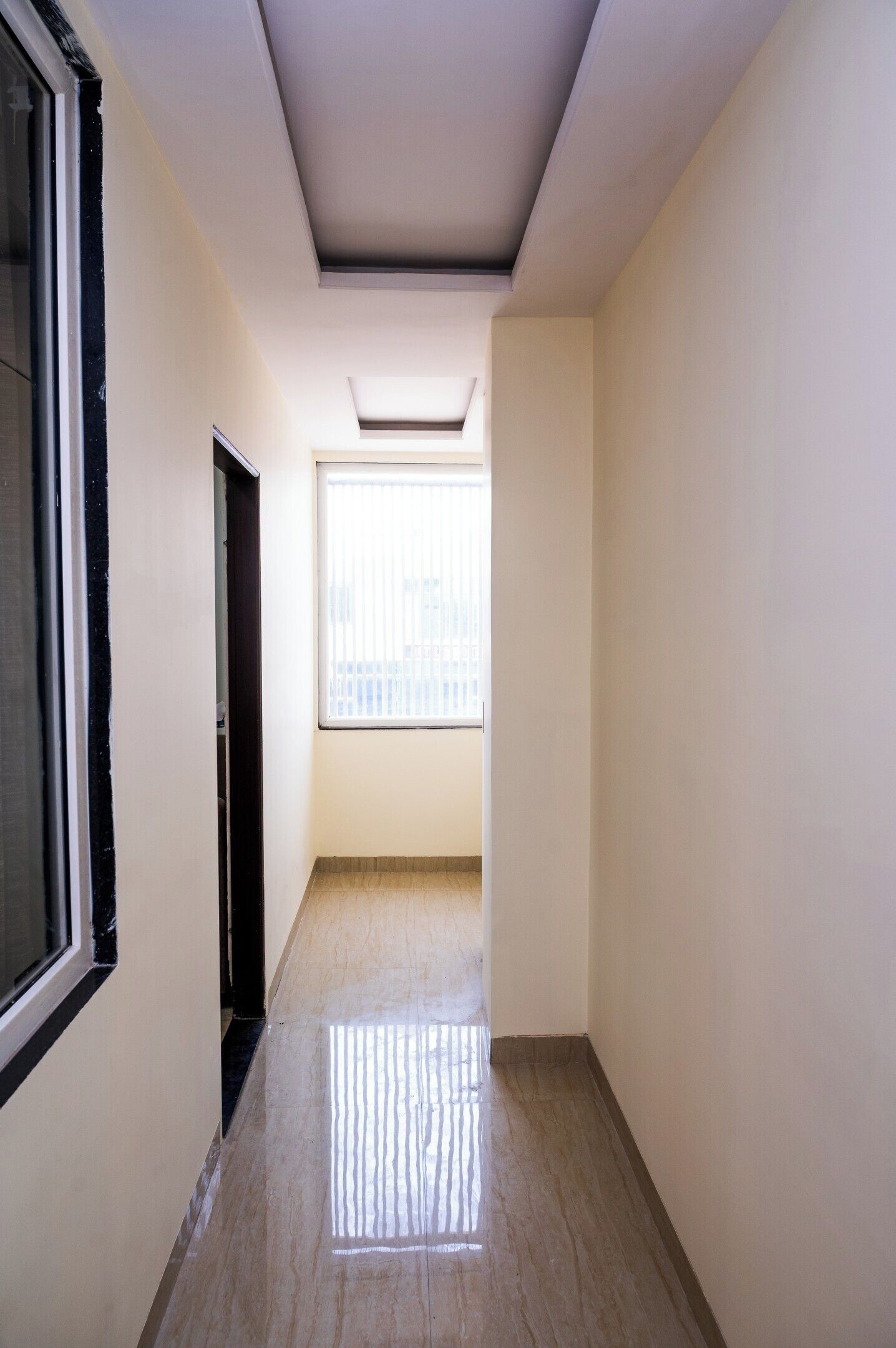 Un pasillo estrecho con suelo de baldosas, una ventana con persianas y una puerta a la izquierda.