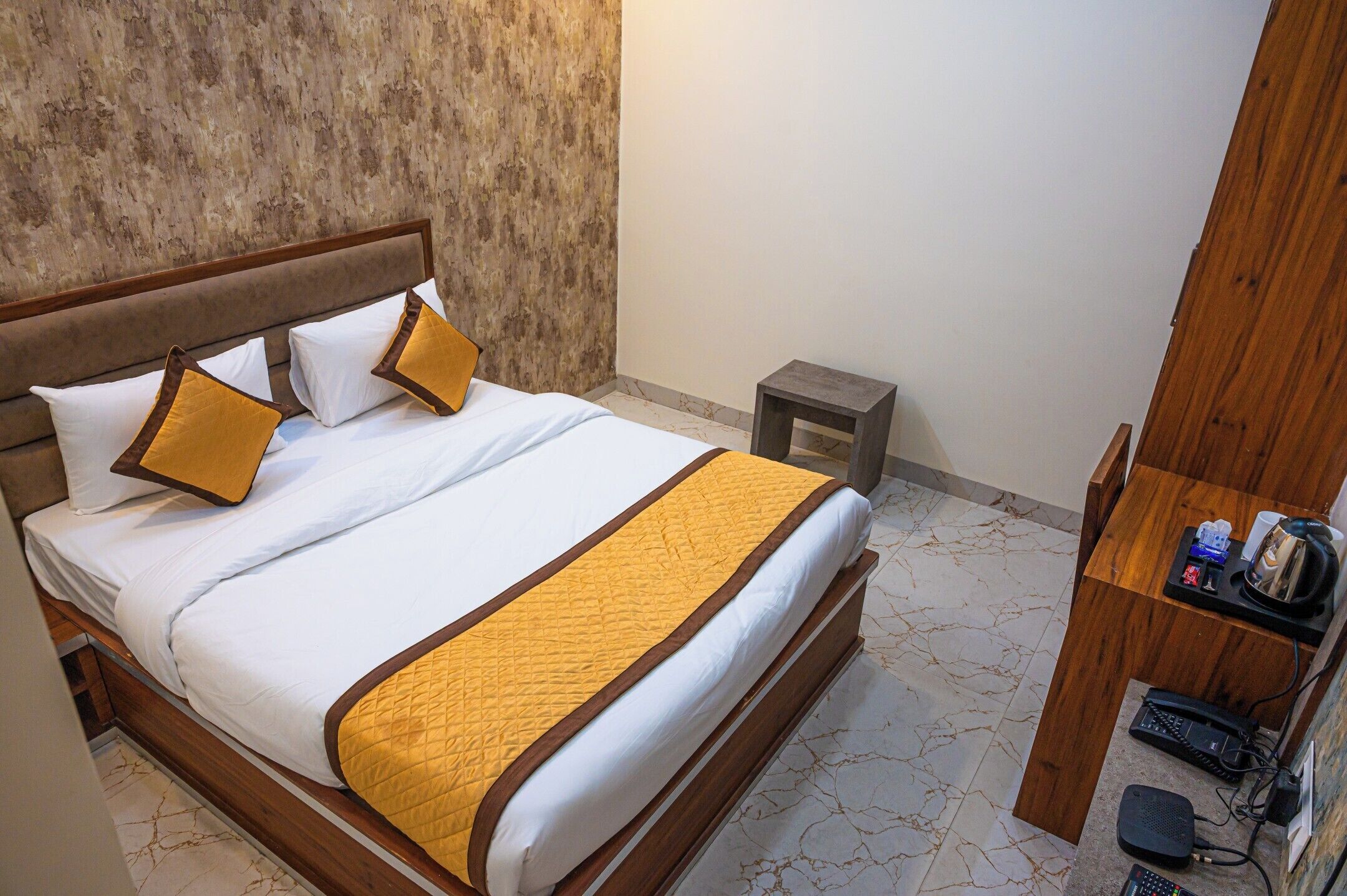 A neatly made bed with white linens and a yellow accent, a small bedside table with a lamp, and a wooden nightstand with a phone and charger.
