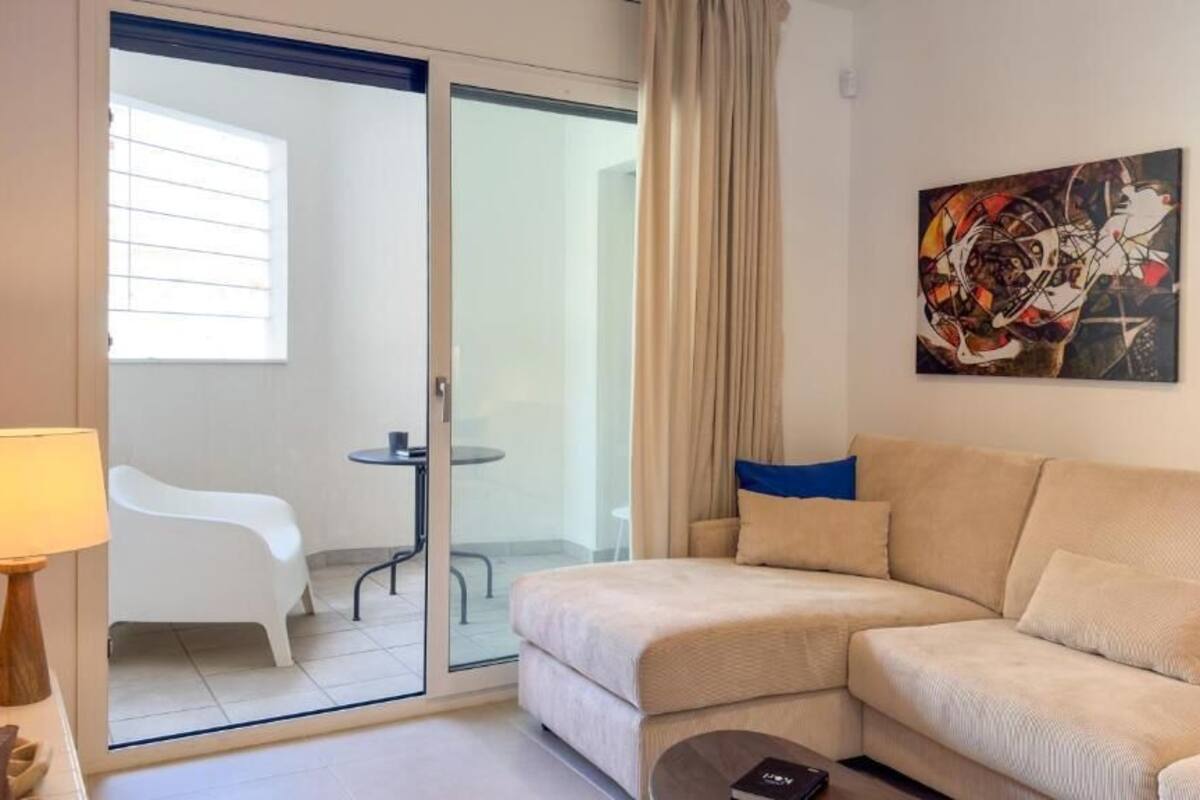 A modern living room with a beige sofa, a glass door leading to a balcony, a small table, and a lamp.
