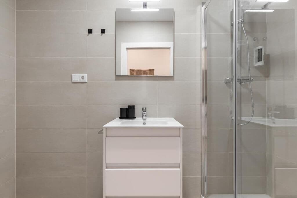 Een moderne badkamer met een glazen douchecabine, een witte wastafel met een zwarte zeepdispenser en een spiegel boven de wastafel.