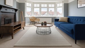 A living room with a fireplace, a blue sofa, a round coffee table, and a view of buildings through large windows. - Beautiful 1 bed flat in Wimbledon (London)