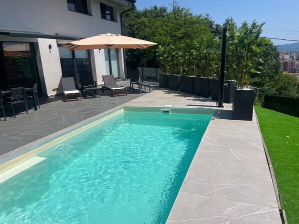 A rectangular swimming pool with clear blue water, surrounded by a tiled patio with lounge chairs and an umbrella.