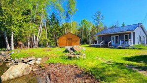 A cabin with a wooden exterior and a metal roof, surrounded by a grassy yard and a small stream. - Southbrook Cottage-Waterfront*Pet Friendly*A/C*Washer/Dryer (Greenville)