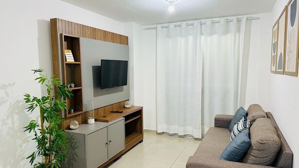 Una sala de estar moderna con un televisor de pantalla plana montado en un mueble de madera, un sofá marrón con cojines, una planta en maceta y un ventanal con cortinas translúcidas.