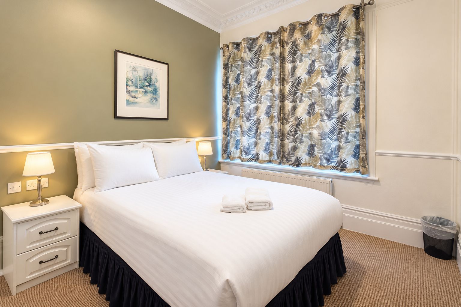 A hotel room with a bed, nightstands, a lamp, a framed picture, a window with curtains, and a radiator.