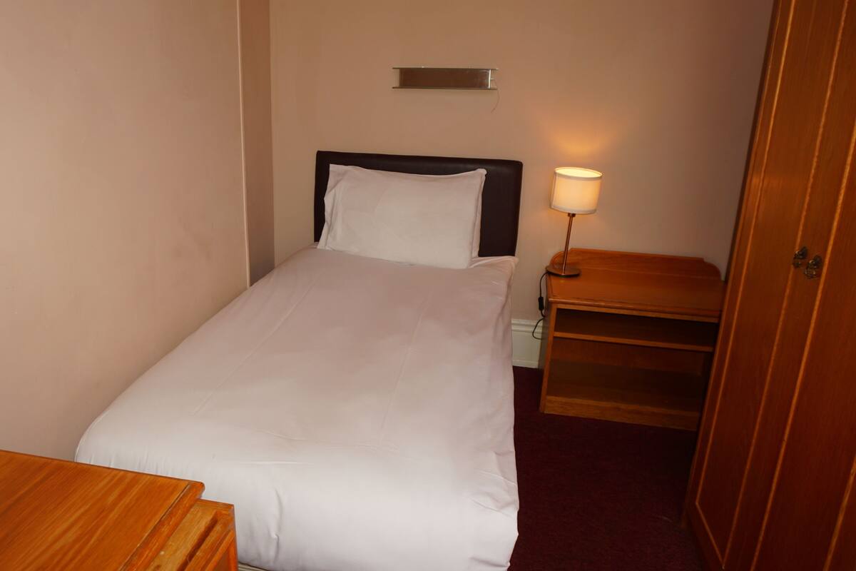 A single bed with a white mattress and pillow, a bedside table with a lamp, and a wooden nightstand.