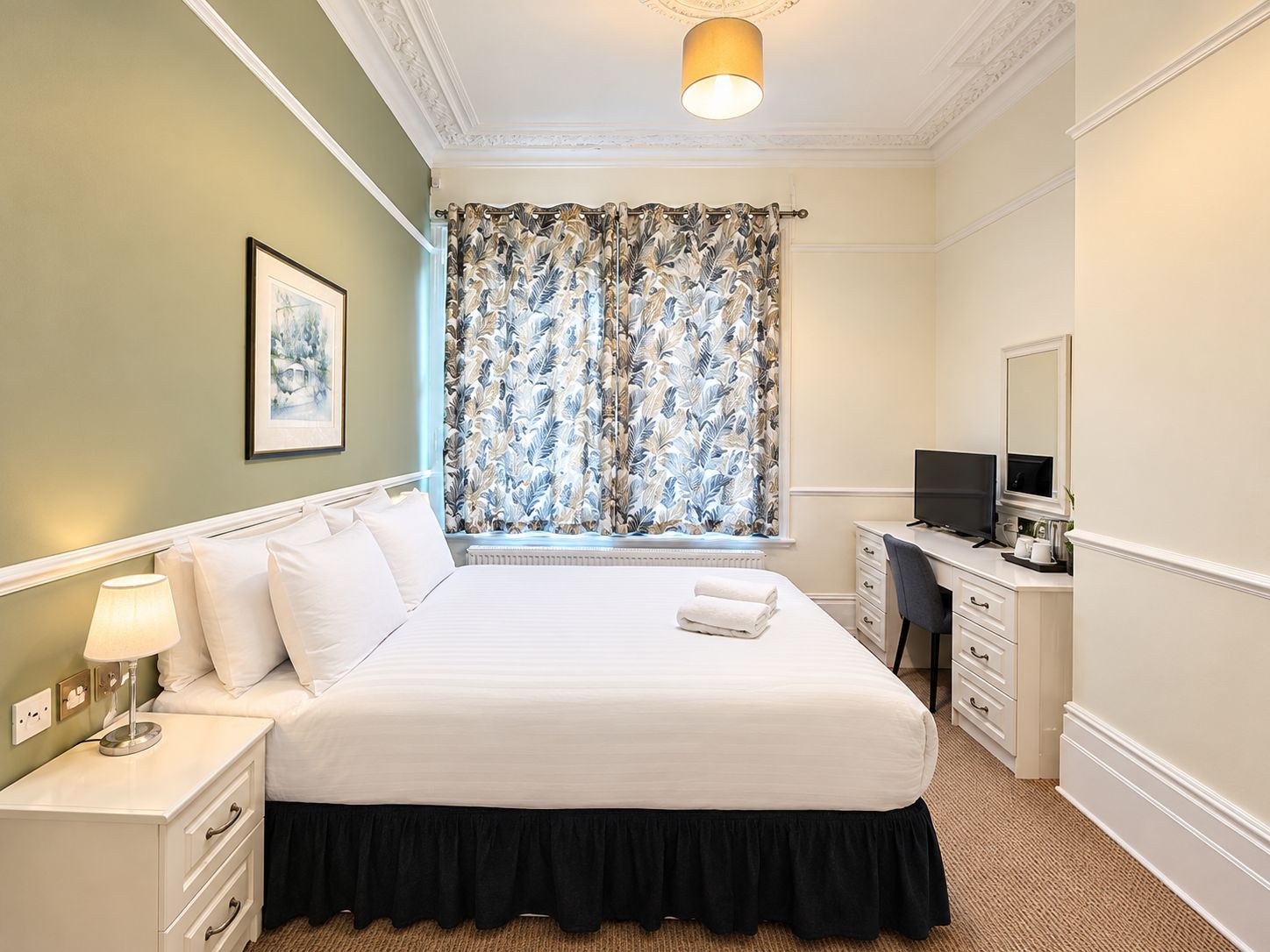 Quarto de hotel com cama, escrivaninha com computador, janela com cortinas, luminária na parede e quadro emoldurado.