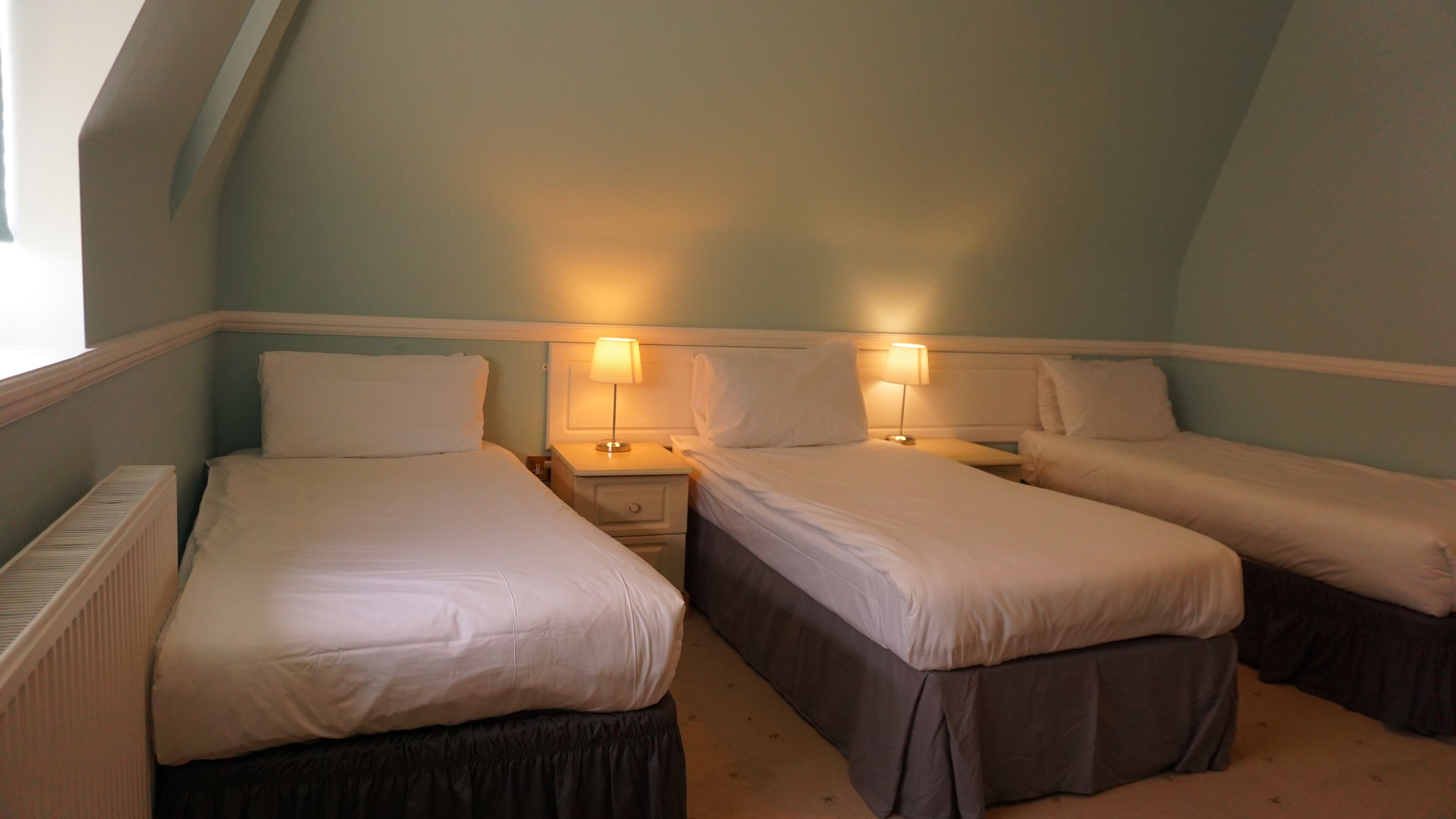 Una habitación con tres camas individuales, cada una con una mesita de noche y lámpara. La habitación tiene techo inclinado y paredes azul claro.
