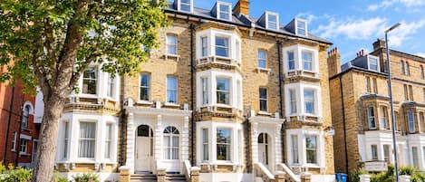 A row of terraced houses with a central entrance, a tree in the foreground, and a clear sky.