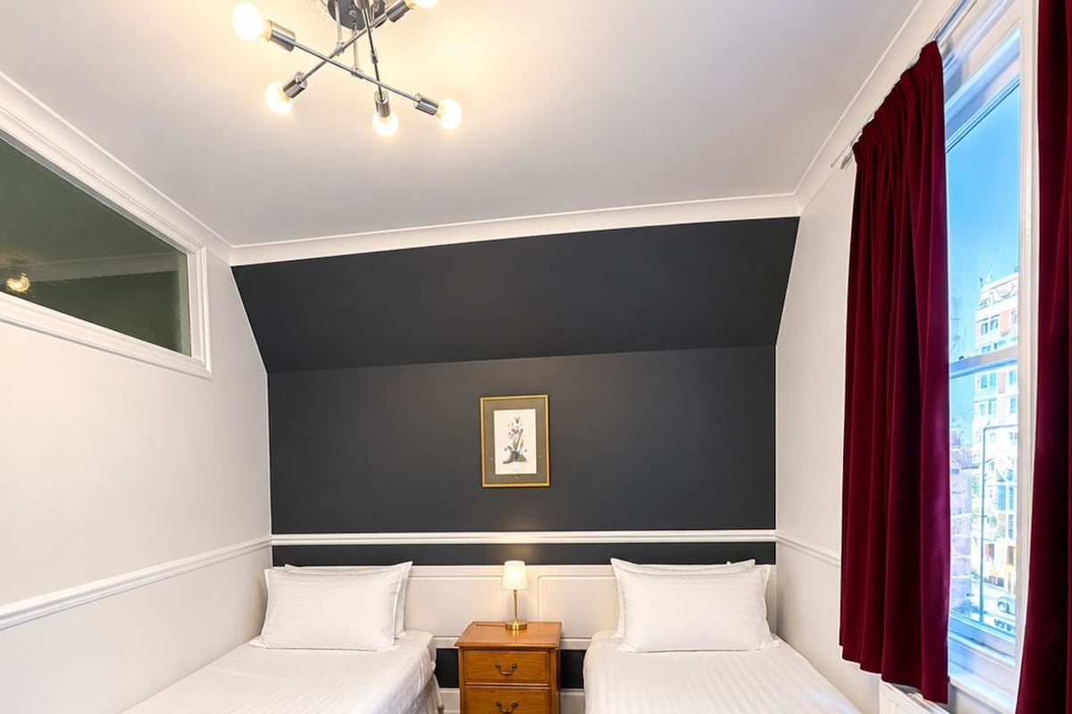 Habitación de hotel con dos camas individuales, una mesita de noche, una lámpara, un cuadro en la pared y una ventana con cortinas.