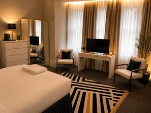 A hotel room with a bed, two chairs, a desk with a TV, a mirror, and a window with curtains. - Albany Hotel (Hove)