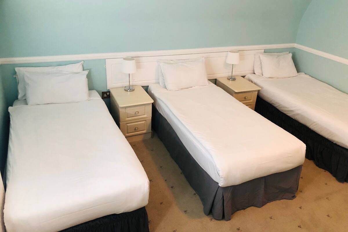 A room with three single beds, each with a bedside table and lamp. The walls are painted light blue, and the room has a simple, functional design.