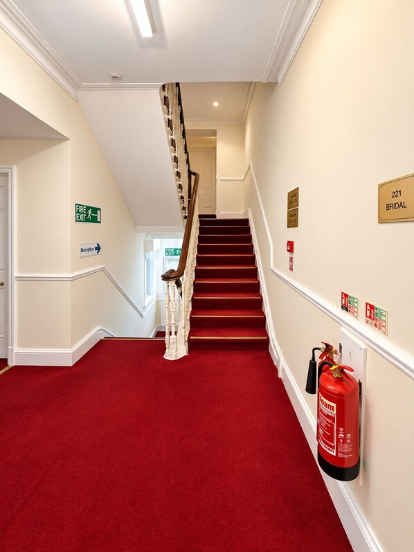 A hotel hallway with a red carpet, a staircase, and a fire exit sign. - Albany Hotel (Hove)