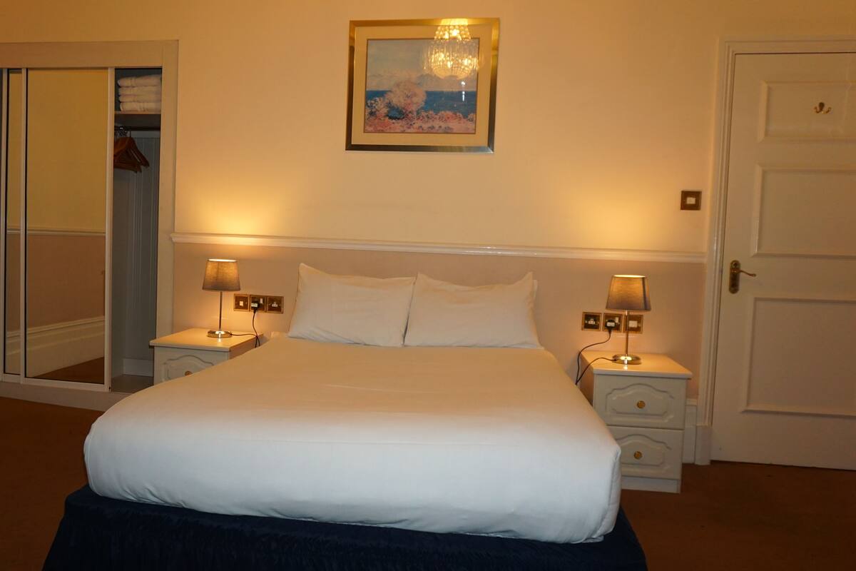 A hotel room with a bed, two bedside tables with lamps, a door, a mirror, and a painting on the wall.