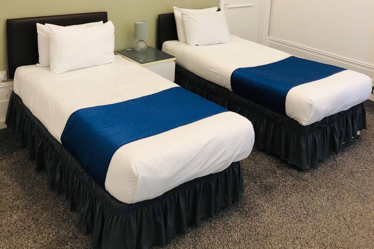 A hotel room with two beds, each with white bedding and blue accents, a nightstand with a lamp, and three wall-mounted flower paintings.