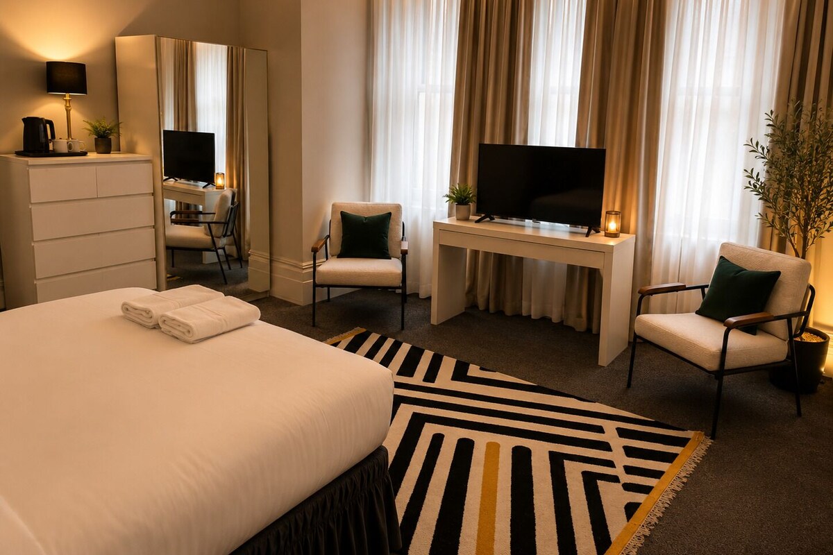 Una habitación de hotel con una cama, un escritorio con televisor, dos sillas, un espejo y una planta.