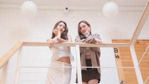 Dos personas de pie en una escalera de madera con barandillas blancas, un candelabro en el techo y una puerta de madera con el número 'B4' a la vista.