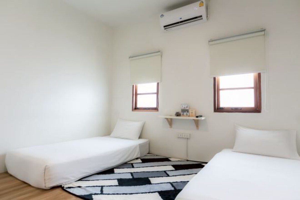 Una habitación pequeña y ordenada con dos camas, un tapete blanco y negro, y una ventana con marco de madera.