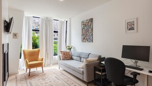A modern living room with a sofa, a chair, a desk with a computer, and a large window with curtains. - Charming 1-bedroom condo in fabulous San Francisco with WiFi, AC fully furnished (San Francisco)
