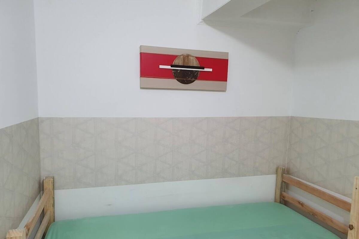 Un lit simple avec un matelas vert, dans une pièce dont un mur est décoré d’un motif géométrique et d’une bande horizontale rouge et beige.