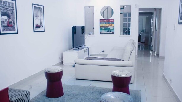 A modern living room with a white sofa, a flat-screen TV, and a coffee table. There are two red stools and a rug on the floor. - WR7 Hostel (Sao Bernardo do Campo)