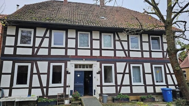 A traditional half-timbered house with a front door, windows, and a small garden area with plants and a bench. - Green apartment on the estate (Nordstemmen)
