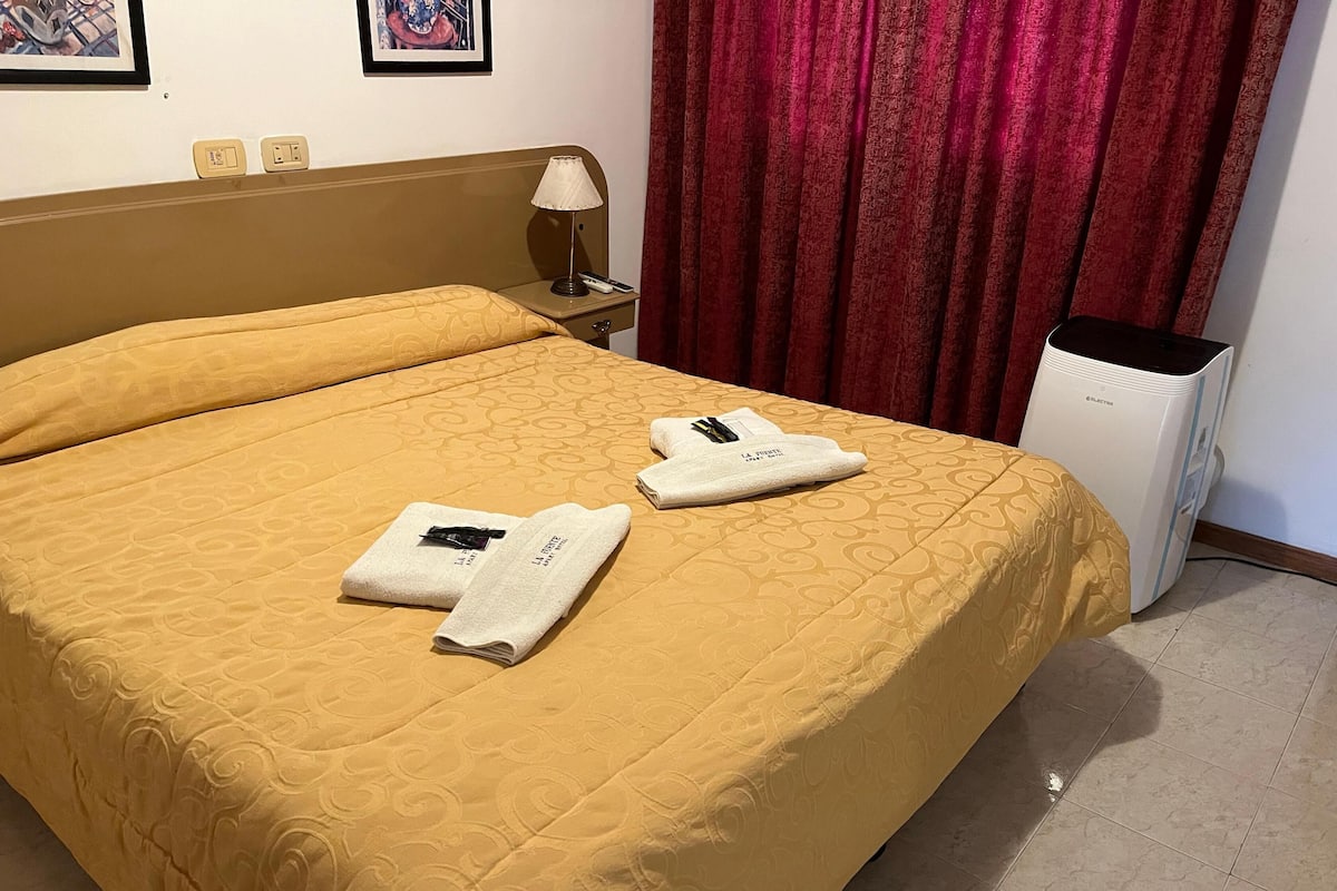 A hotel room with a large bed, two towels on the bed, a nightstand with a lamp, a red curtain, and a heater.