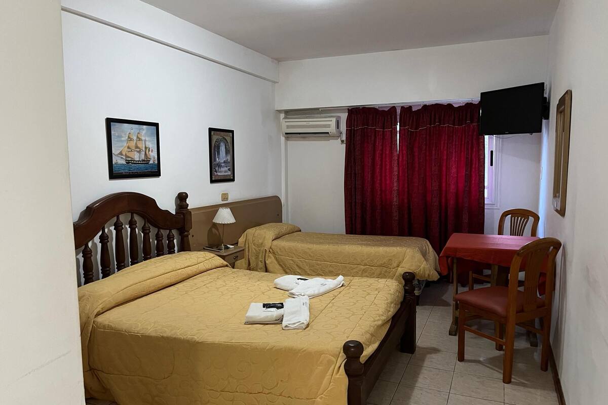 A hotel room with two beds, a table with a chair, a TV, and a window with red curtains.