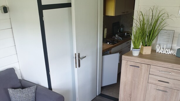 Een knus woongedeelte met een grijze bank, een houten kast en een keukenruimte zichtbaar door een open deur.