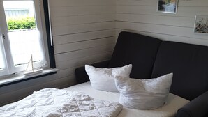 Een eenpersoonsbed met een gewatteerde witte deken, twee witte kussens en een zwart hoofdbord. Een klein raam met uitzicht op groen is zichtbaar.