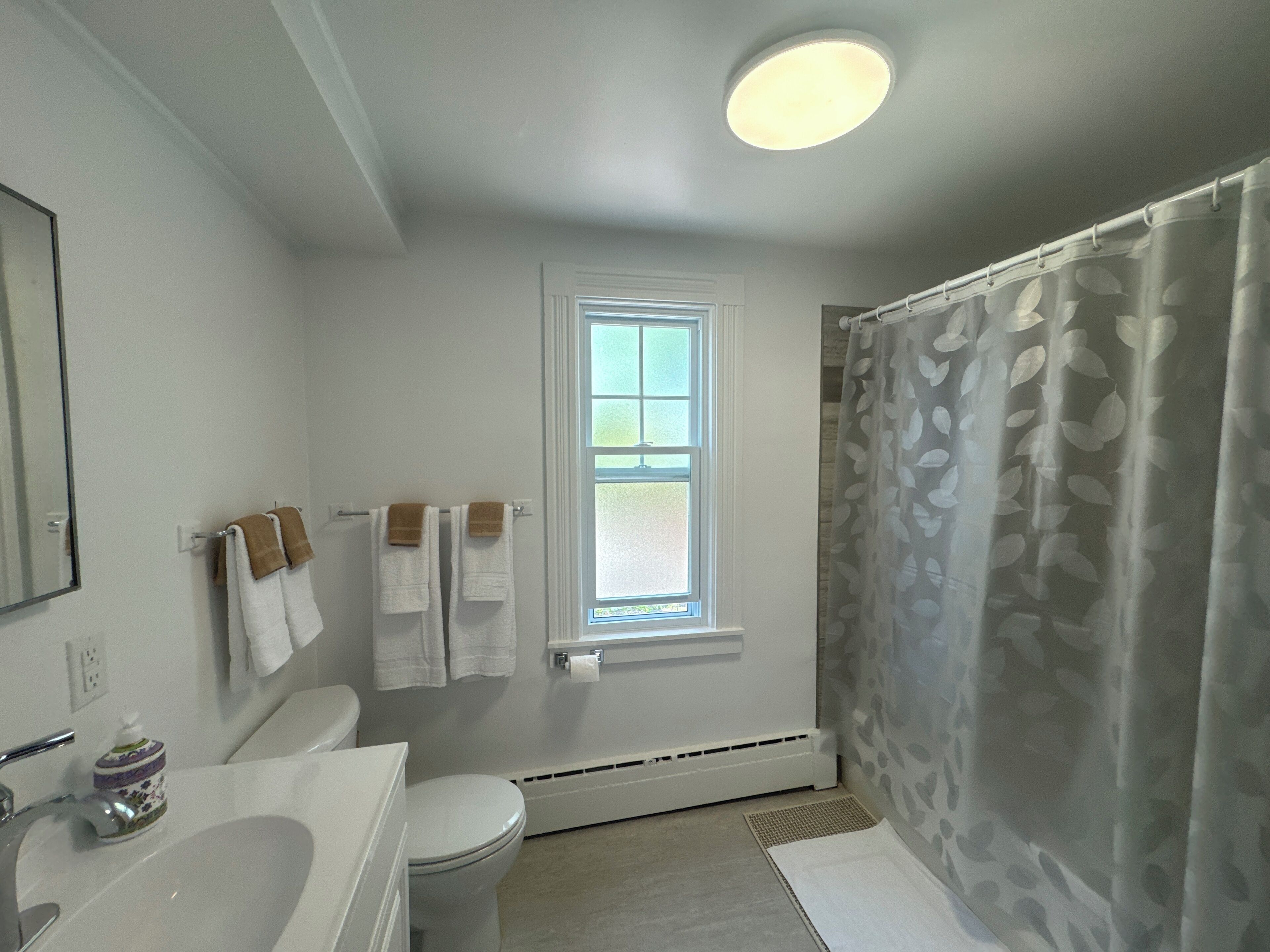 Une salle de bain avec un lavabo blanc, des toilettes et un rideau de douche. Il y a une fenêtre au-dessus des toilettes et un porte-serviettes avec des serviettes.