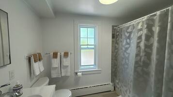Une salle de bain avec un lavabo blanc, des toilettes et un rideau de douche. Il y a une fenêtre au-dessus des toilettes et un porte-serviettes avec des serviettes.