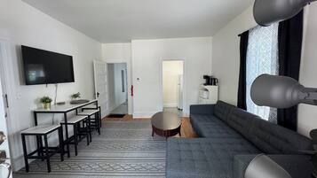 A modern living room with a grey sofa, a dining area with a table and chairs, a flat-screen TV mounted on the wall, and a coffee machine on a white cabinet.