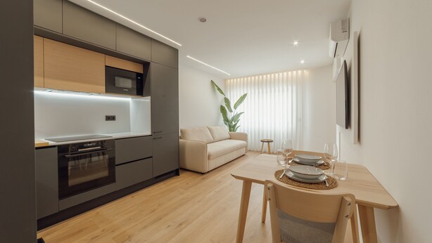 Een moderne keuken met inbouwapparatuur, een eethoek met een houten tafel en een groot raam met gordijnen.