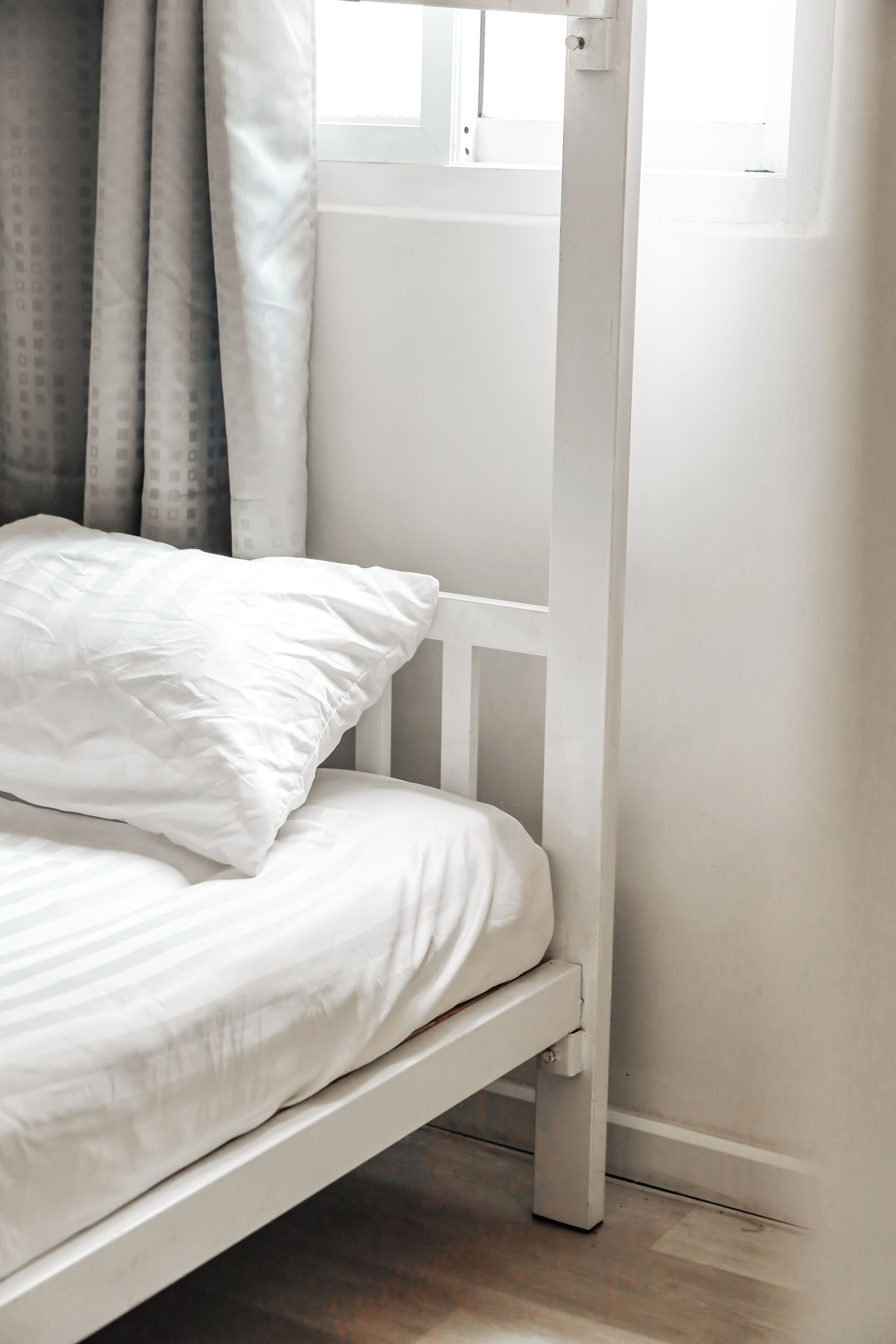 A bed with white bedding and a wooden frame, a window with curtains, and a wooden floor.