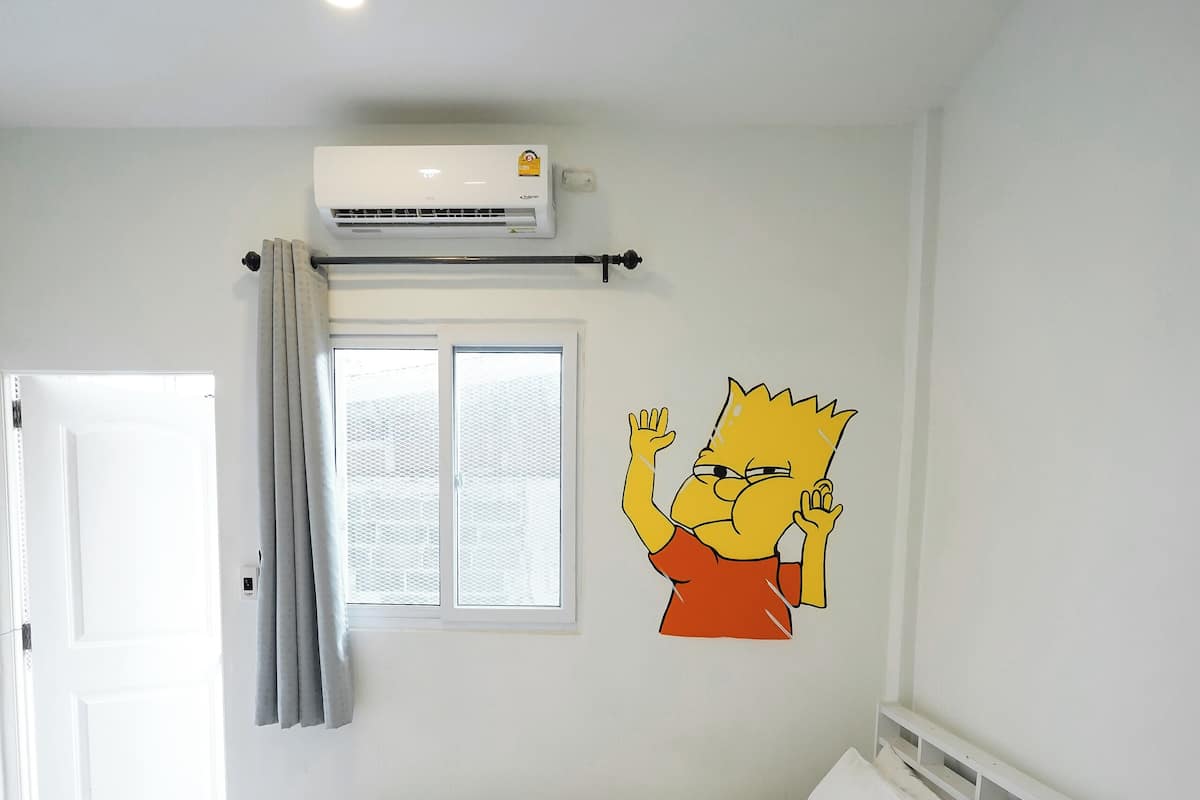 A neatly made bed with white bedding, a wall-mounted air conditioner, a window with curtains, and a cartoon character decal on the wall.