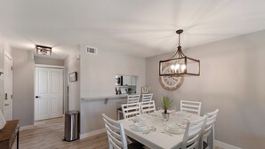 A dining area with a white table set for six, surrounded by white chairs. A large wall clock and a pendant light are visible. - Gulf View Getaway |  Sand & Sunset Views Daily! (Holmes Beach)