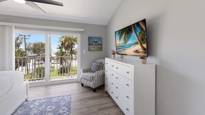 A bedroom with a balcony, a bed, a dresser, a chair, and a painting of a beach scene. - Gulf View Getaway |  Sand & Sunset Views Daily! (Holmes Beach)