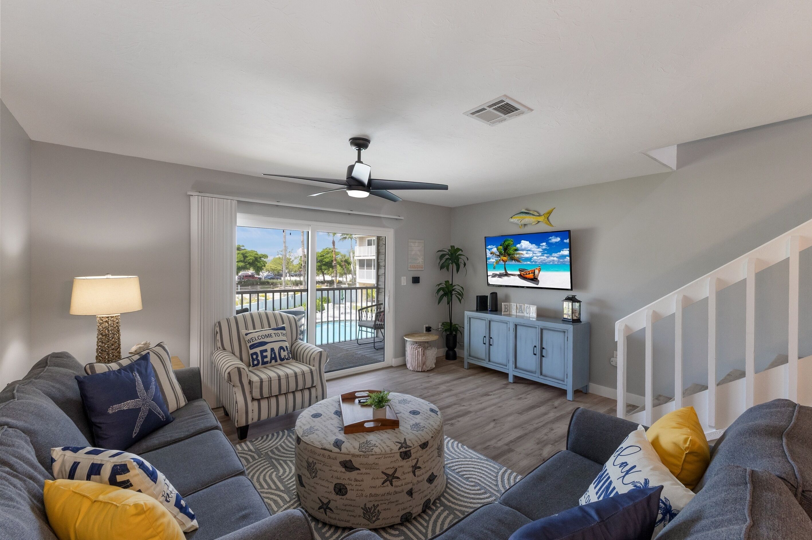 Ein Wohnzimmer mit grauem Sofa, Sesseln mit Streifenmuster und Hocker, einem Deckenventilator, einem Fernseher und Blick auf einen Pool durch eine Schiebetür.