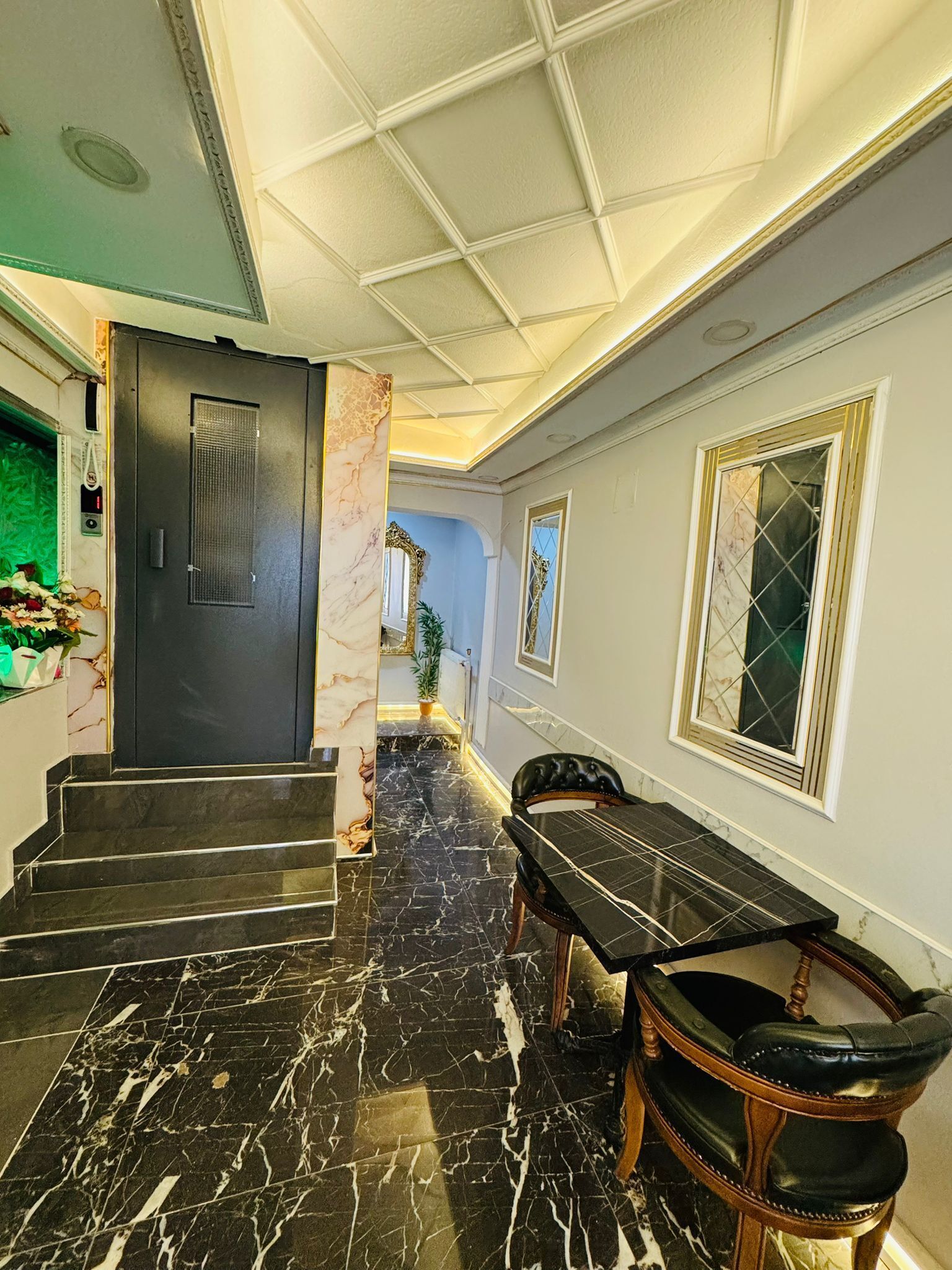 A hallway with marble flooring, a black door, a dining table with chairs, and a mirror on the wall.