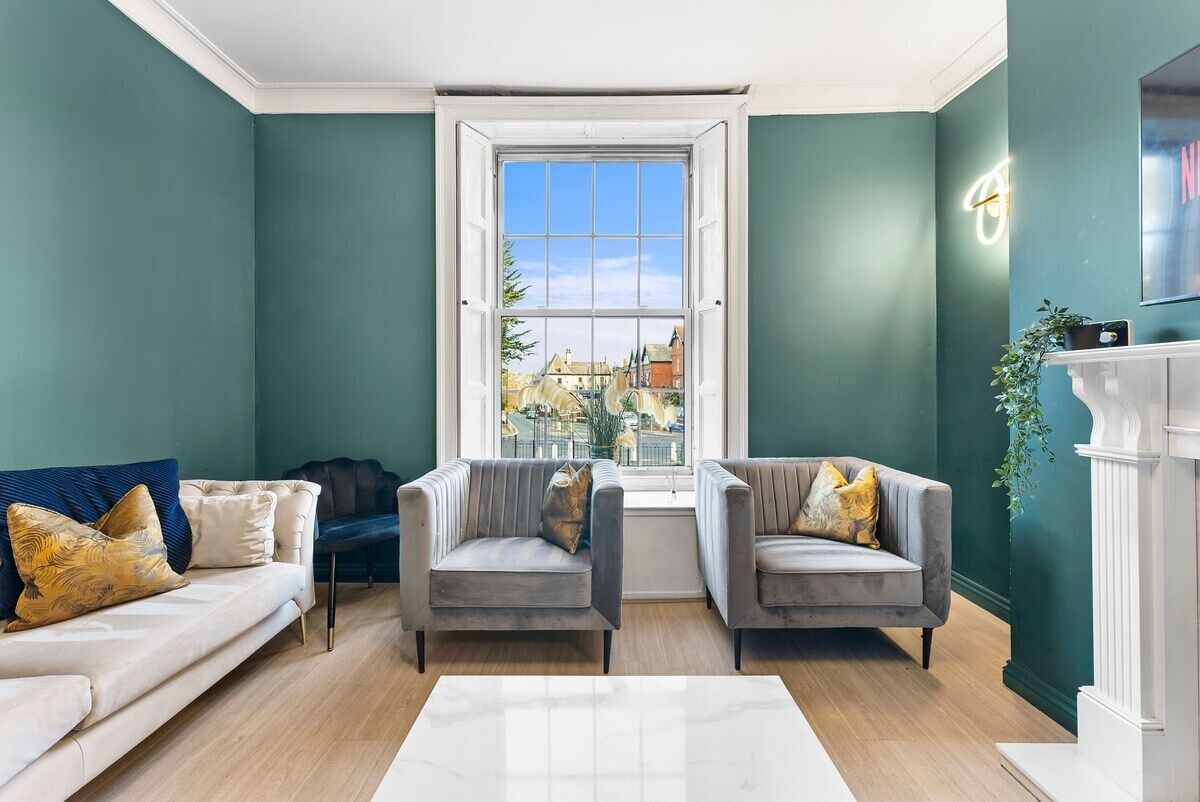A living room with teal walls, a white fireplace, and a large window offering a view of buildings outside.