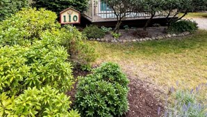 Una casa de dos pisos con techo verde y revestimiento color beige, rodeada de abundante vegetación y un pequeño jardín.