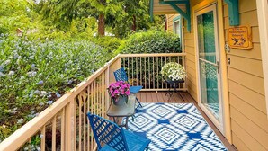Una terraza de madera con un tapete azul estampado, una silla de metal y una maceta con flores rosadas.
