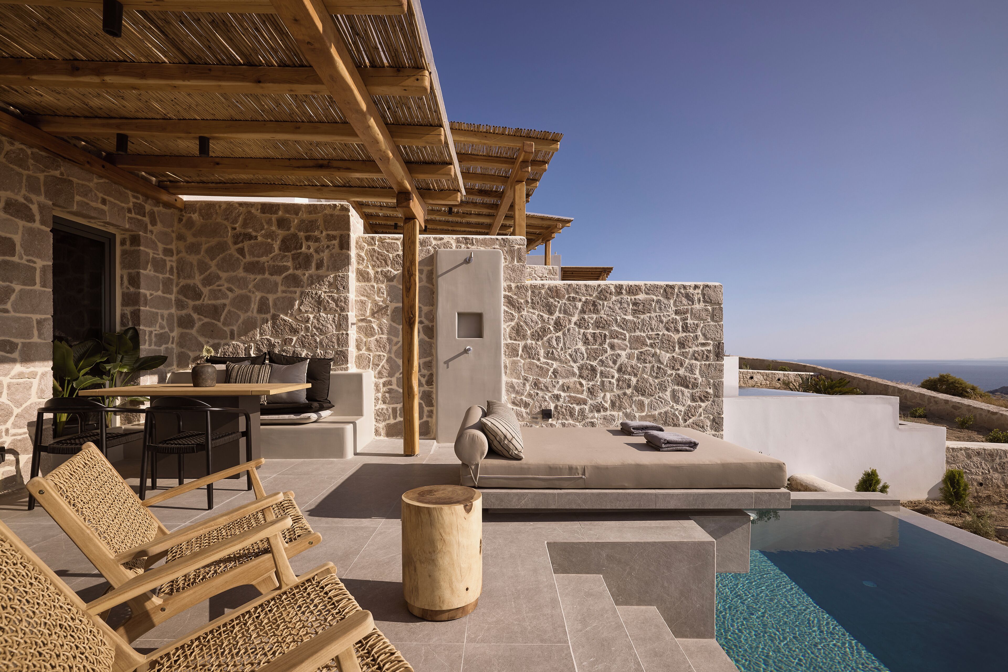 A stone-walled outdoor area with a pool, wooden furniture, and a view of the sea.