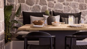A dining area with a stone wall, a wooden table set with a bowl of fruit, a vase, and a pitcher of water. - Kemelia Suites (Milos)
