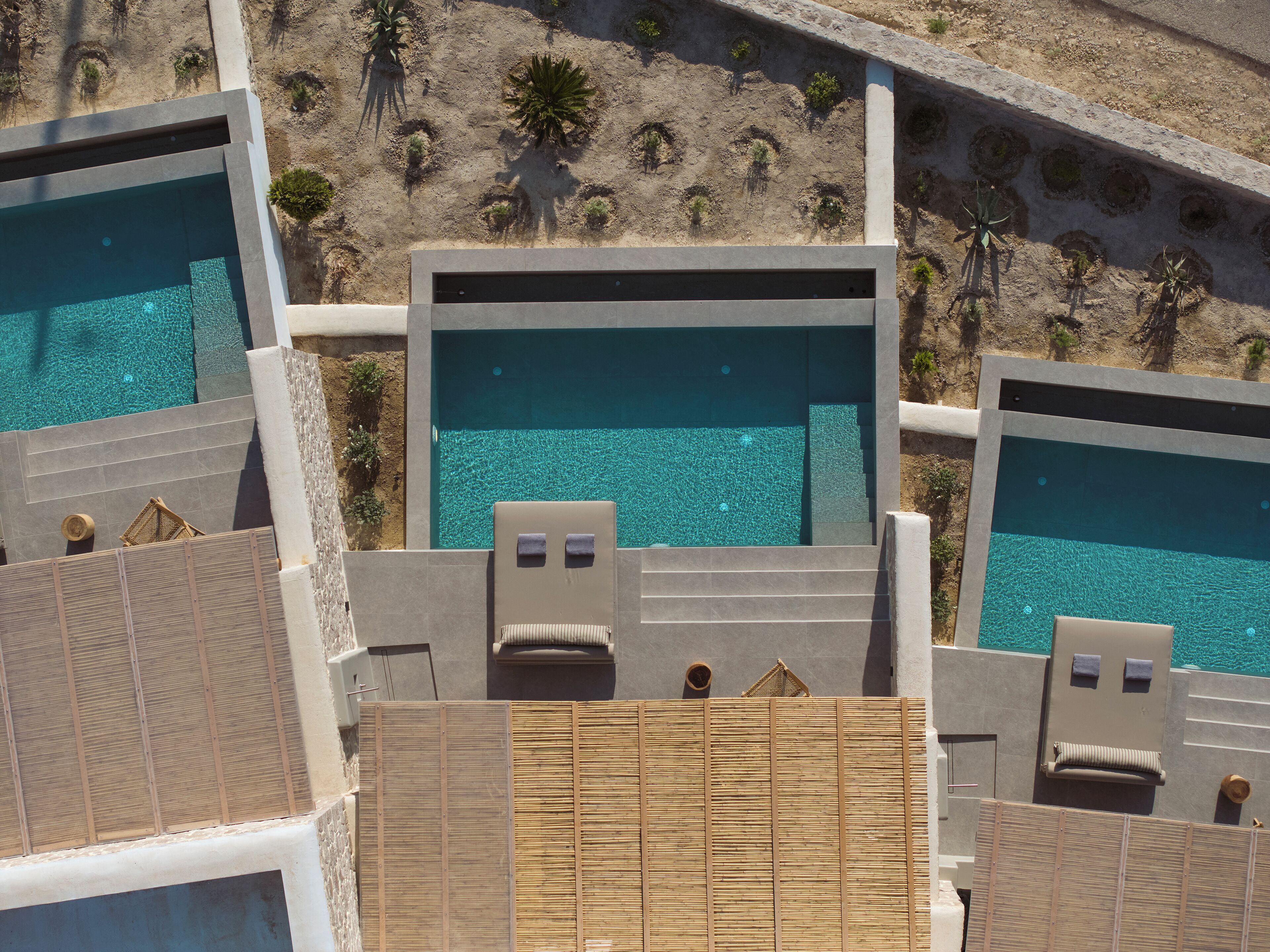 Eine Vogelperspektive zeigt einen luxuriösen Urlaubsort mit mehreren Schwimmbecken, Liegestühlen und einer sandigen Landschaft mit wenig Vegetation.