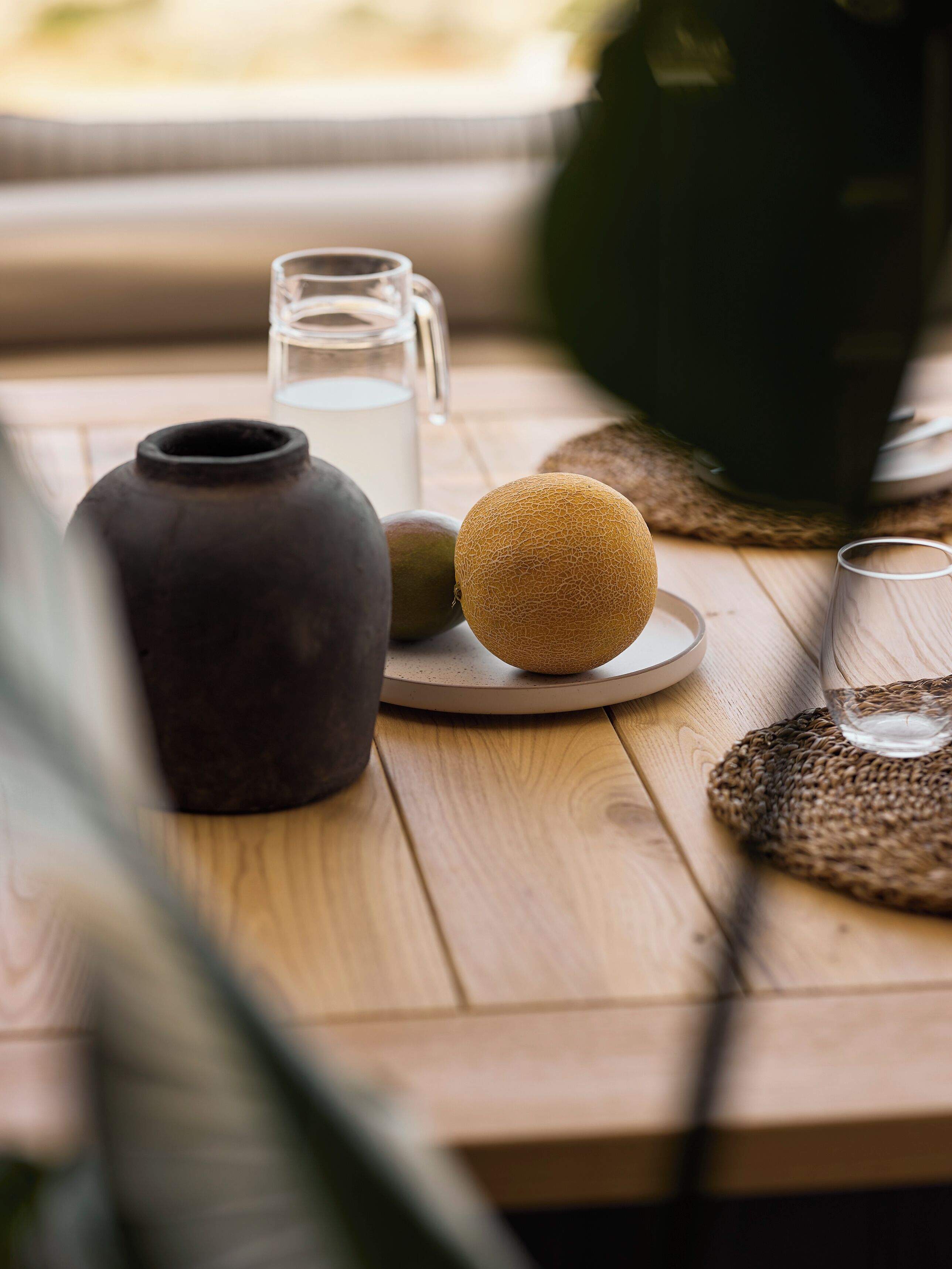 Une table en bois, sur laquelle se trouvent un vase en céramique, une carafe en verre, un verre et un set de table en fibres tressées.