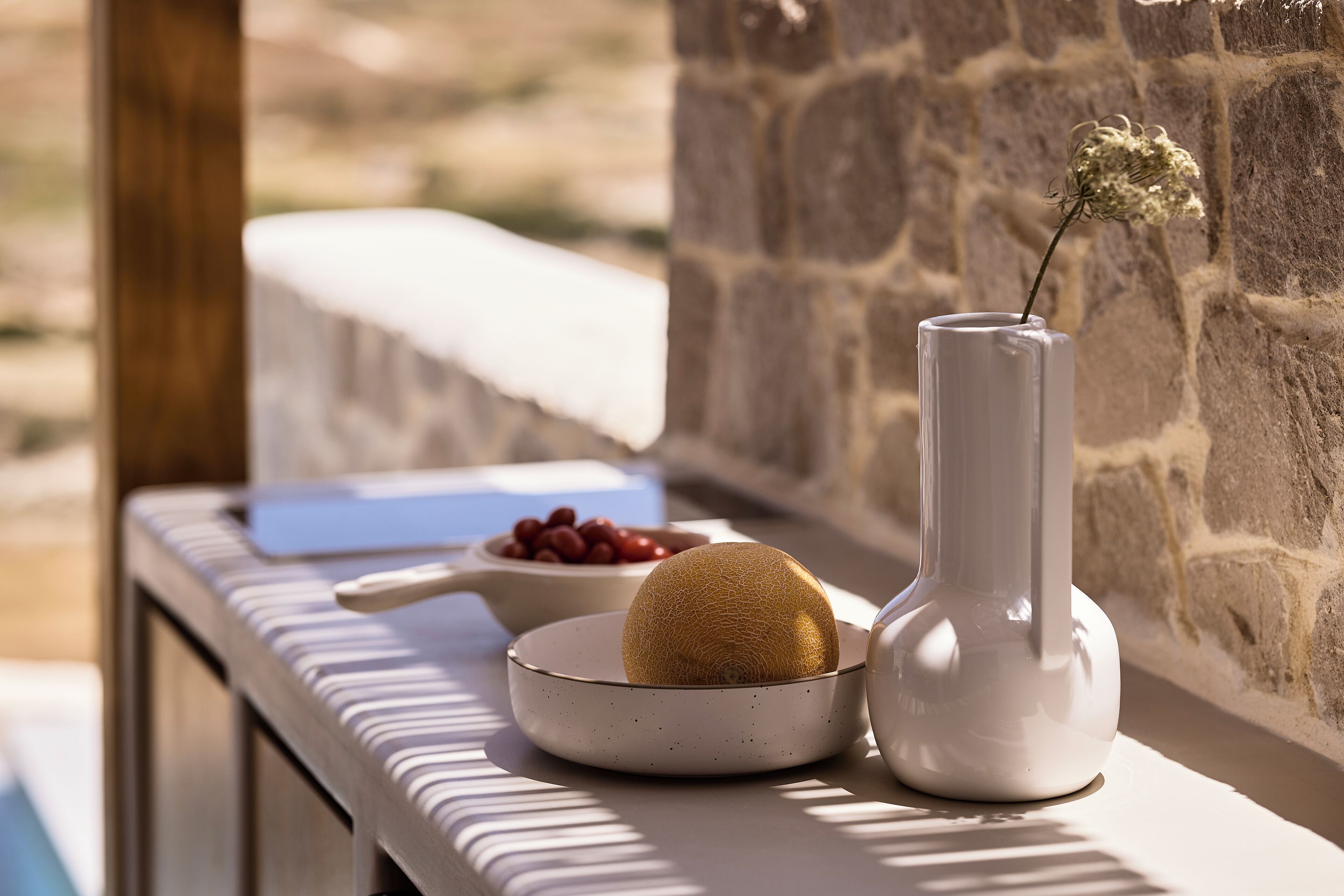 A stone wall, a white vase with a dried flower, a bowl of red berries, and a melon on a table.